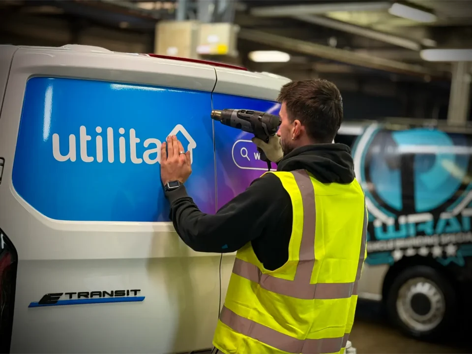 Technician applying blue and pink gradient vinyl branding to Utilita Ford Transit van inside workshop.
